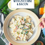 Hands eating a bowl of super cheesy pasta with zucchini and bacon and text title overlay