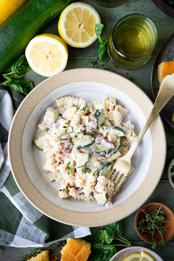 Overhead image of a bowl of cheesy pasta with zucchini and bacon on a green table