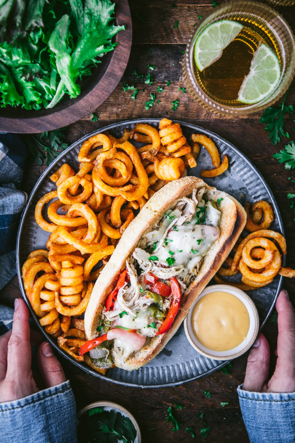 Overhead shot of hands holding a metal plate with a philly chicken cheesesteak with a side of fries
