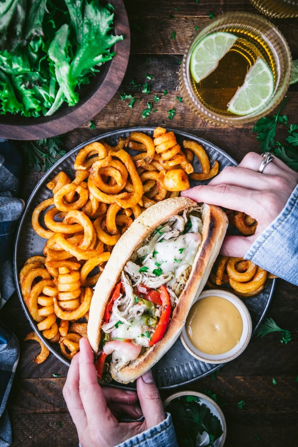 Hands holding a chicken philly cheesesteak on a plate with a side of fries