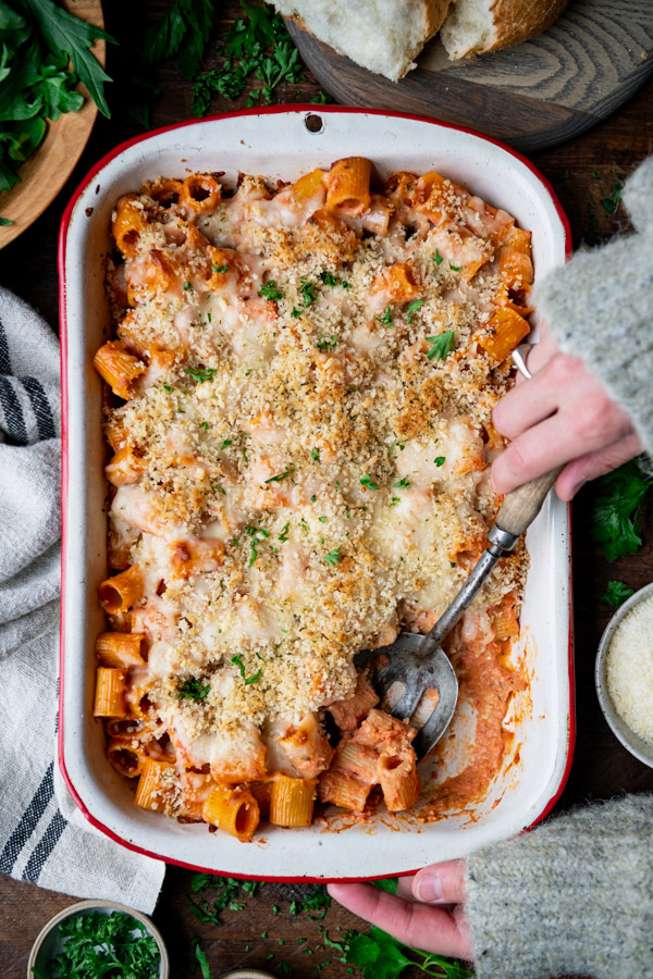 Overhead shot of hands serving baked rigatoni from a casserole dish on a wooden table