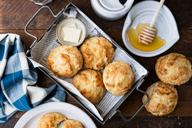 Overhead shot of a basket of buttermilk drop biscuits on a wooden table with honey and jam