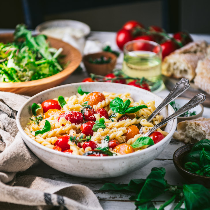 Square side shot of a bowl of pasta with cherry tomatoes and fresh basil on a table with salad and bread in the background