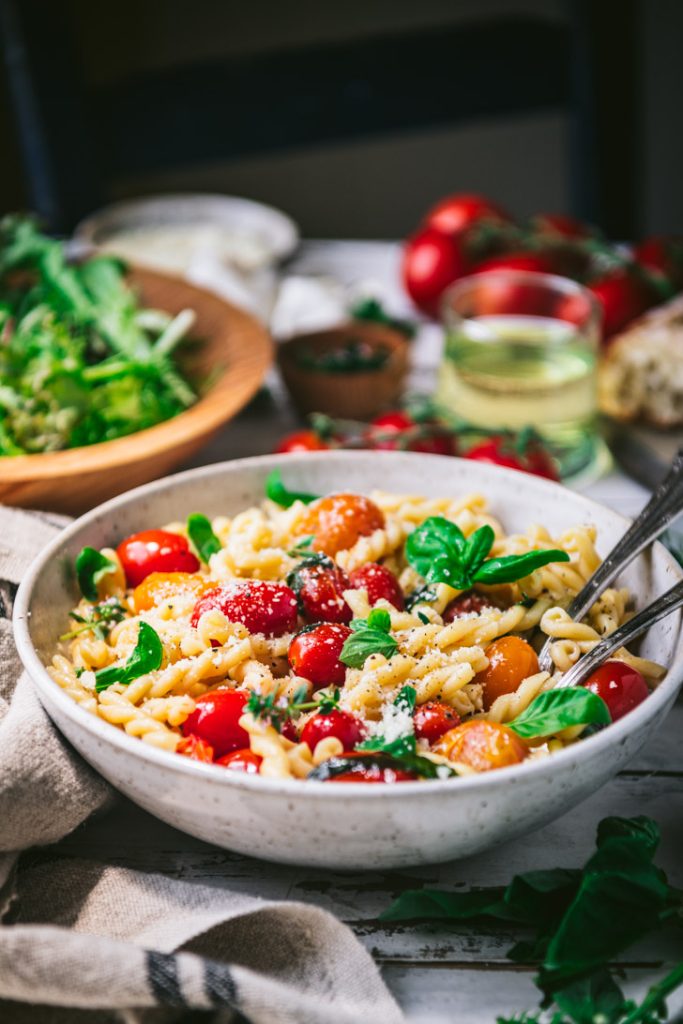 Close up side shot of a bowl of pasta with cherry tomatoes on a dinner table with salad and bread