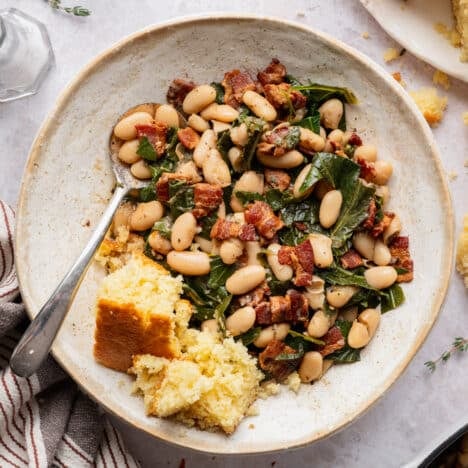 Square overhead image of a spoon in a bowl of beans and greens with cornbread on the side.