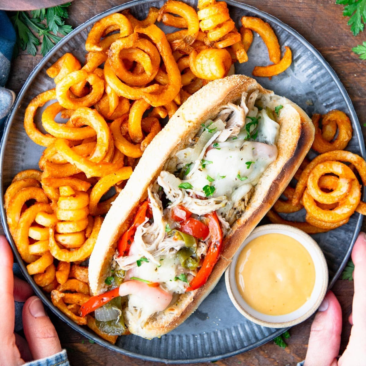 Overhead image of hands holding a plate of chicken Philly cheesesteak with a side of curly fries.