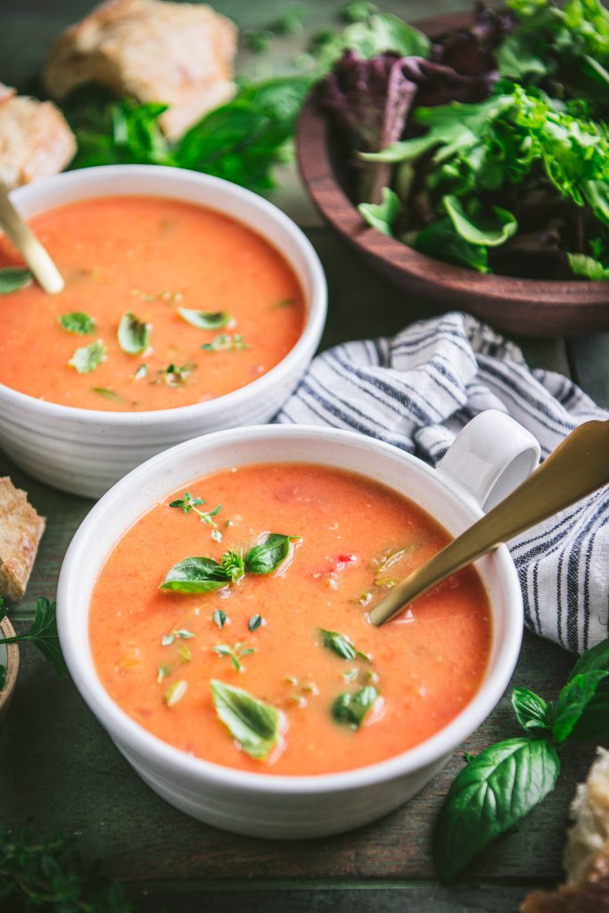 Front shot of two bowls of roasted tomato soup on a green table with a side salad in the background
