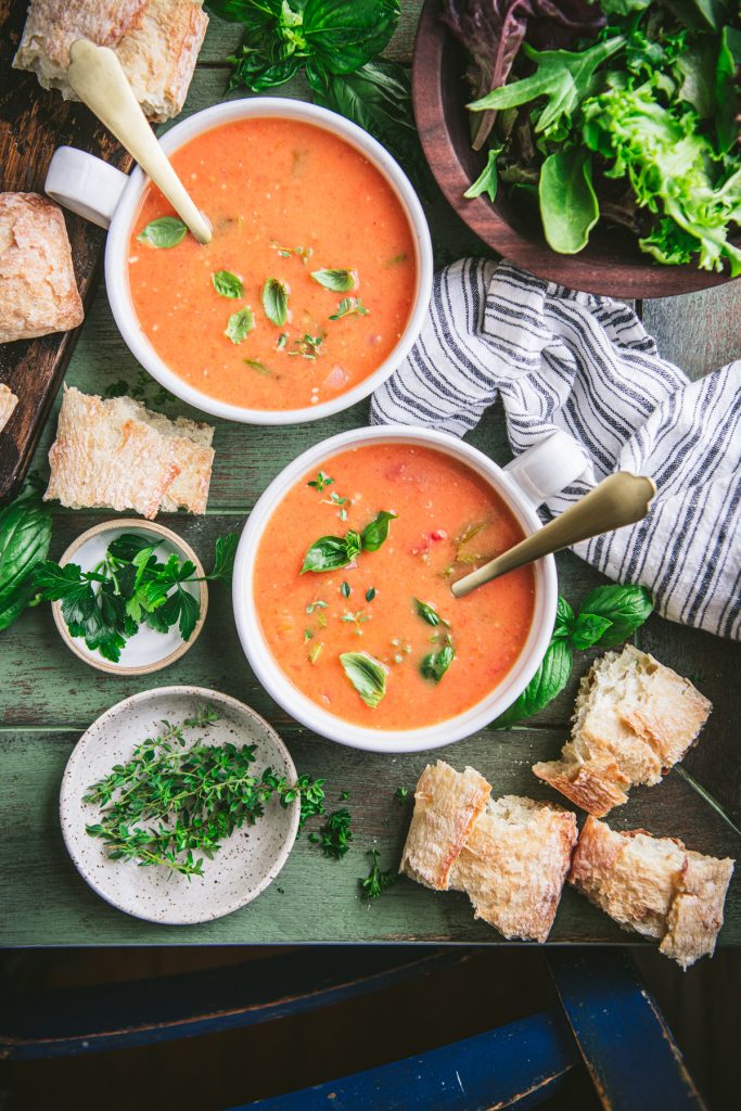 Two bowls of roasted tomato soup on a green table with crusty bread on the side