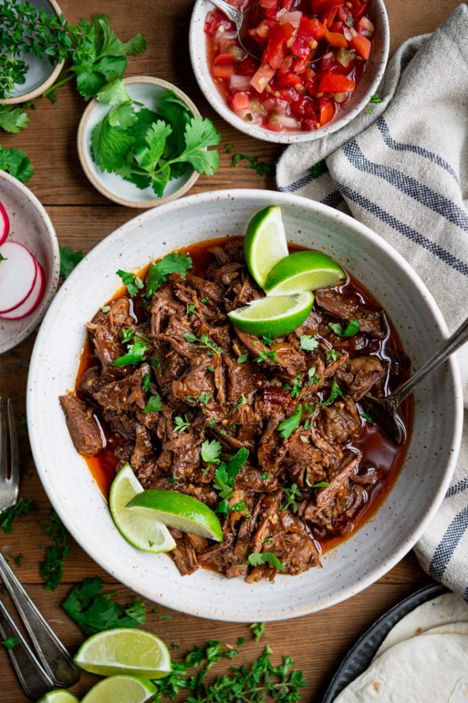Overhead image of a bowl of beef barbacoa with lime wedges and cilantro