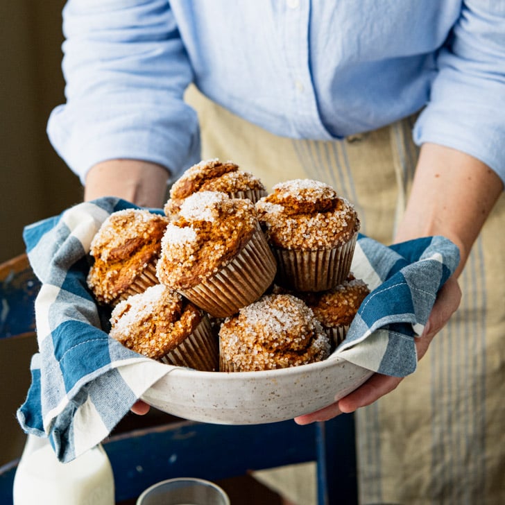 A woman holds a bowl full of freshly baked homemade pumpkin muffins topped with sanding sugar.