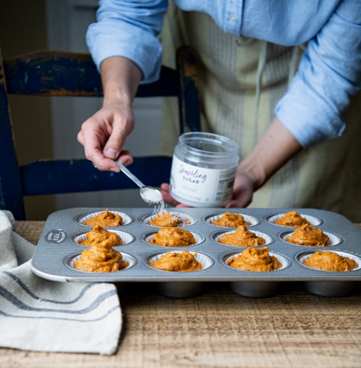 A woman sprinkles crystal sanding sugar on top of unbaked muffins in a metal muffin tin.