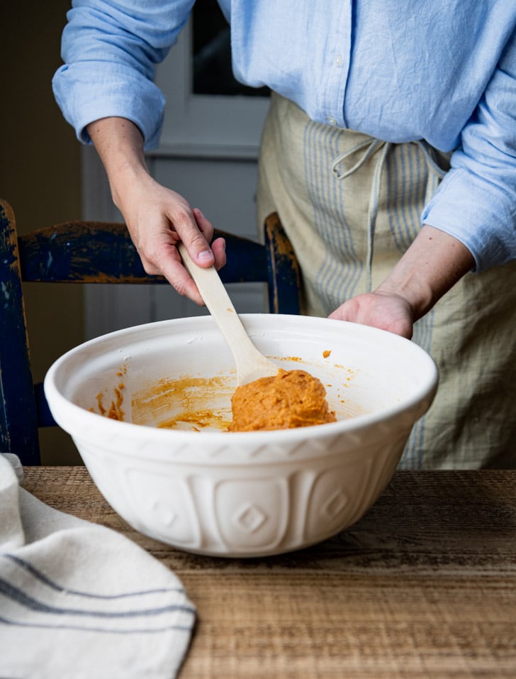 A woman uses a wooden spoon to mix pumpkin muffin batter in a white bowl.