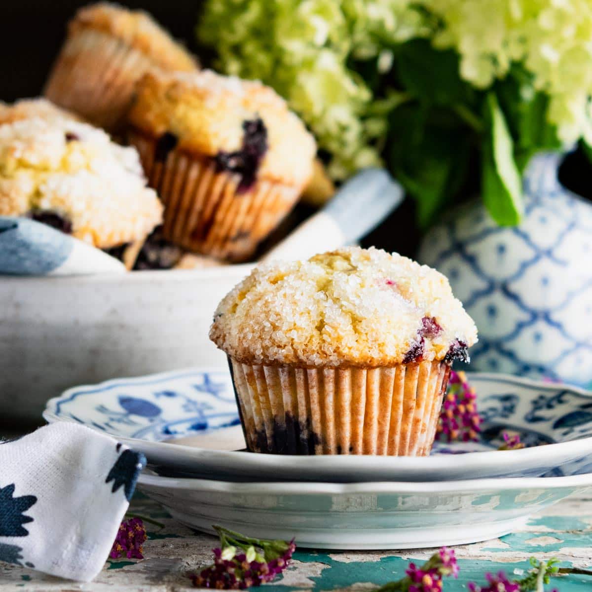 Square side shot of sour cream blueberry muffins on a table with flowers in the background.