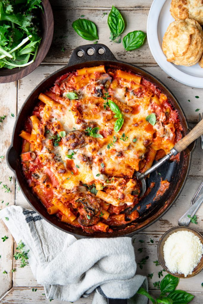Skillet of vegetable baked ziti on a wooden table with fresh herbs