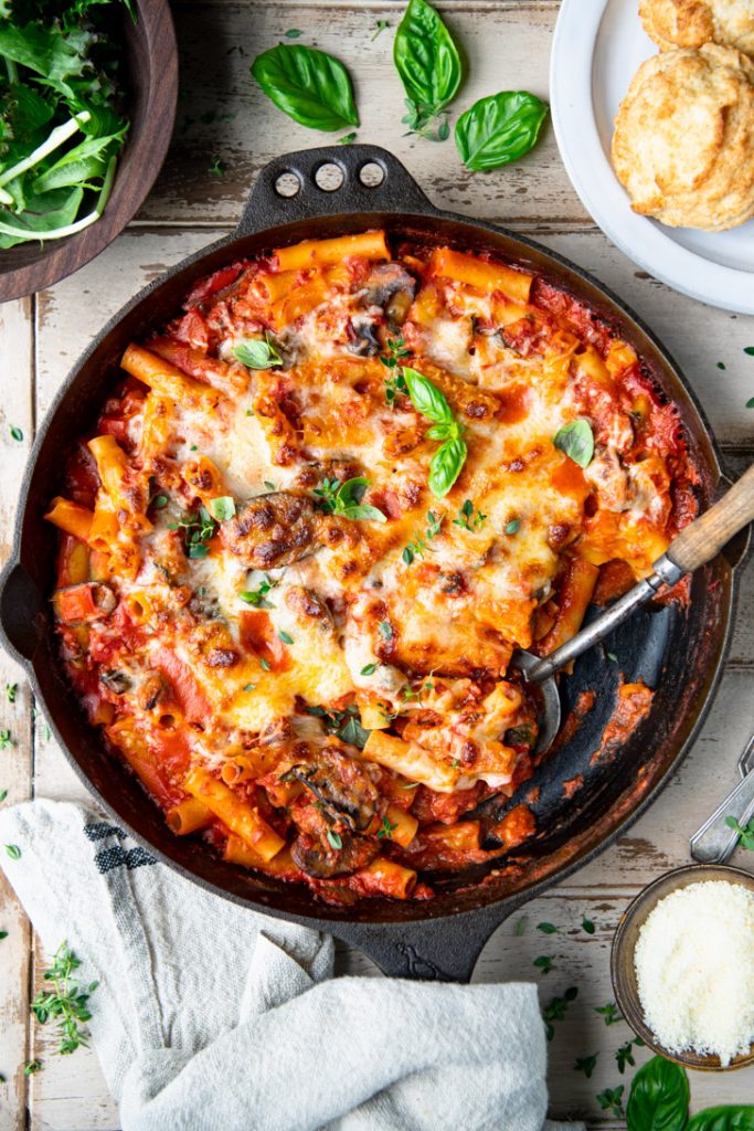 Overhead shot of a pan of vegetable and five cheese baked ziti on a table with salad and bread