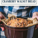 Side shot of hands holding a dutch oven with cranberry walnut no knead bread and text title box at top