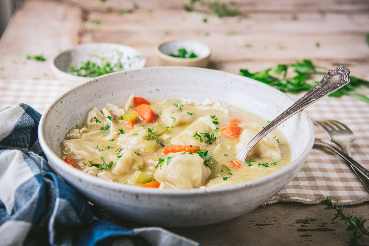 A bowl of chicken and dumplings sits on a dining table along a spread of fresh herbs.