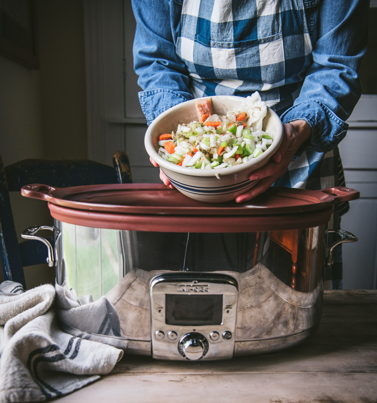 A woman cumps a large bowl of chopped vegetables - onions, carrots, and celery - into a large crockpot.