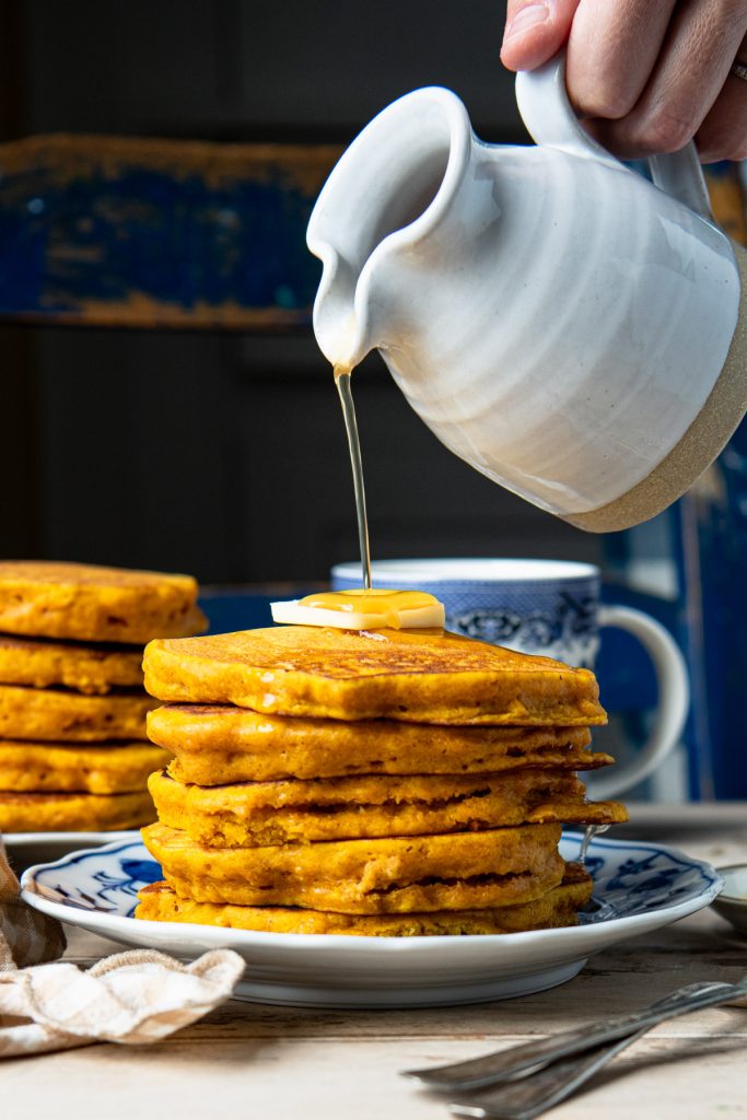 Front shot of a white pitcher drizzling syrup on a stack of pumpkin pancakes
