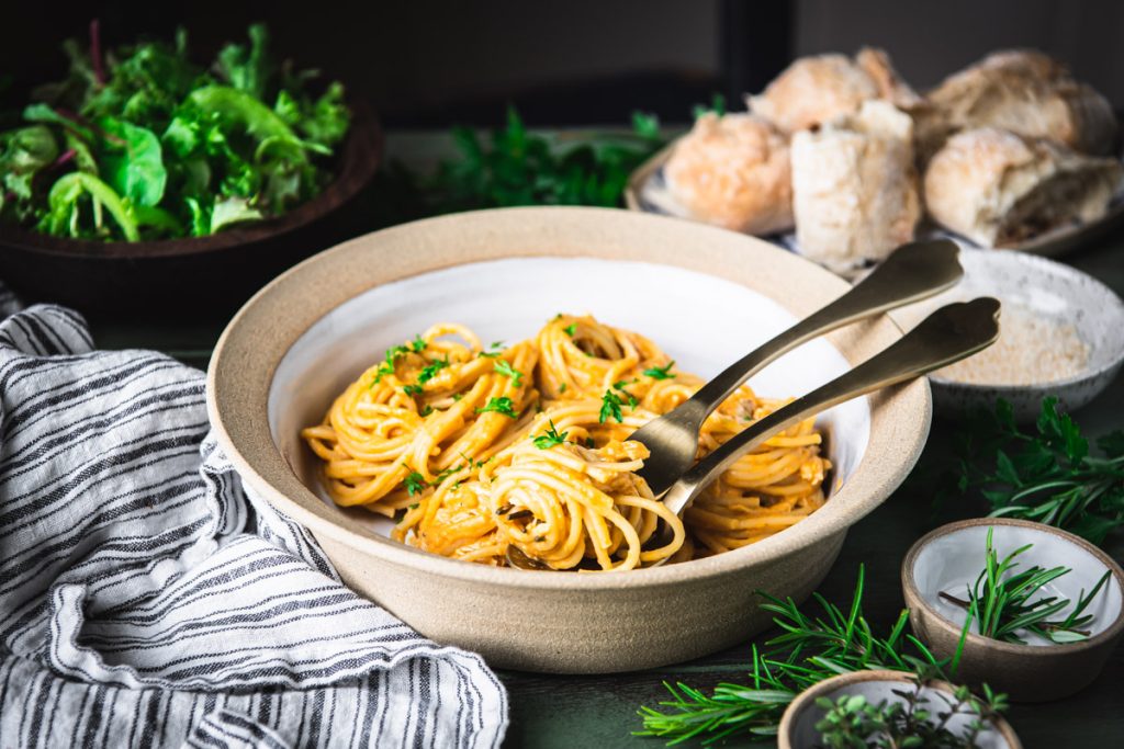 Horizontal image of a bowl of pumpkin pasta with salad and bread in the background