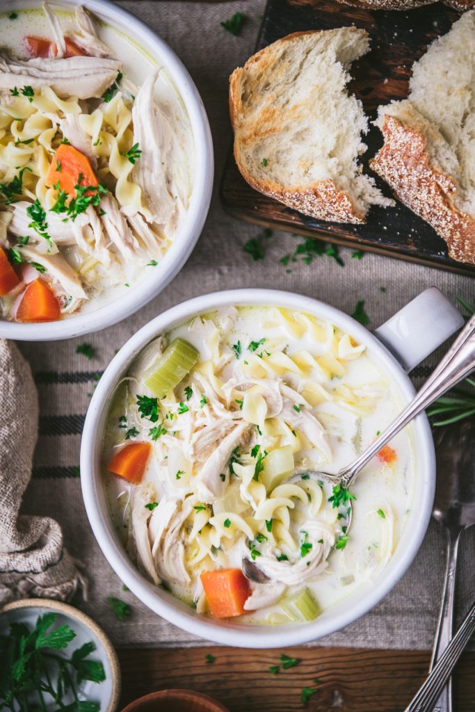 Overhead shot of two bowls of creamy chicken noodle soup on a dinner table with a side of bread