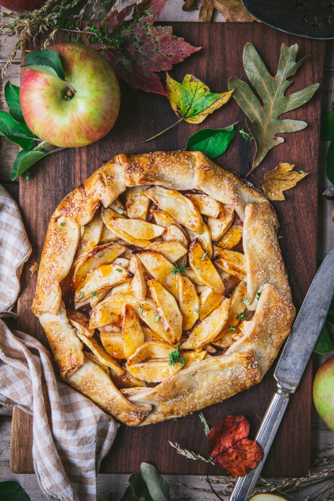 Overhead shot of a rustic apple galette on a wooden cutting board surrounded by fall leaves and apples