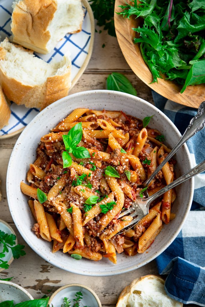 Bowl of one pot pasta with ground beef, salad, and bread on a table