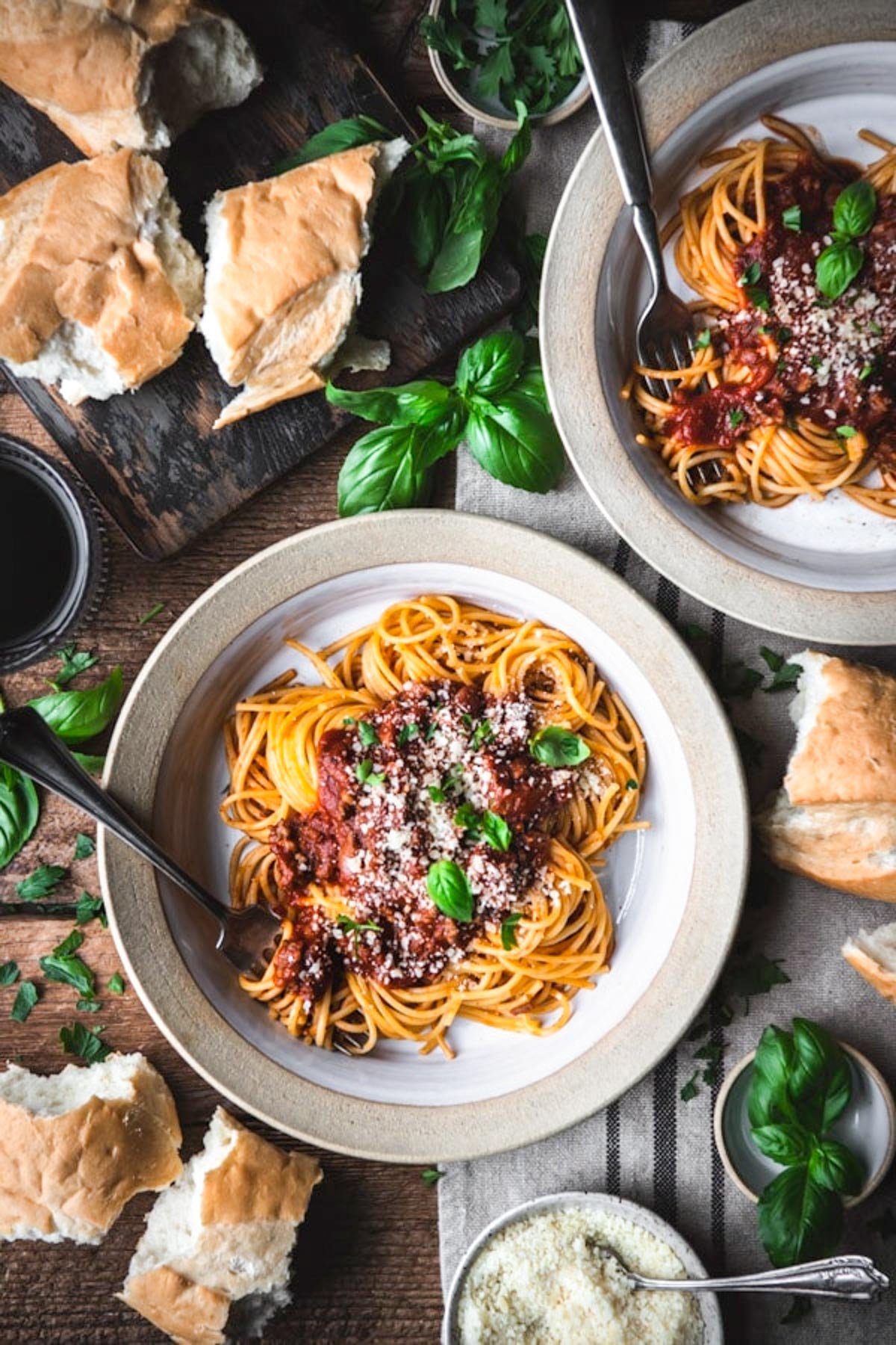 An overhead image of two bowls of spaghetti topped with a homemade spaghetti sauce and parmesan cheese. Pieces of bread, basil leaves, and bowls of grated parmesan cheese surround the bowls.