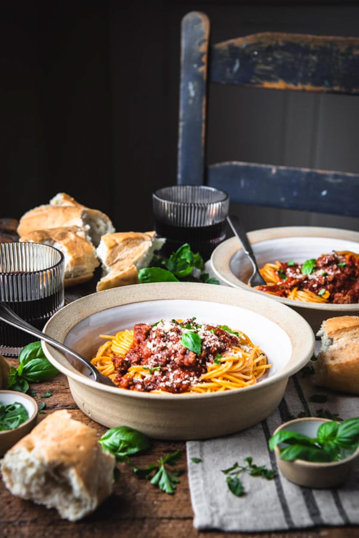 Bowls of homemade spaghetti served at a table surrounded by pieces of french bread, basil leaves, and a glass of red wine.