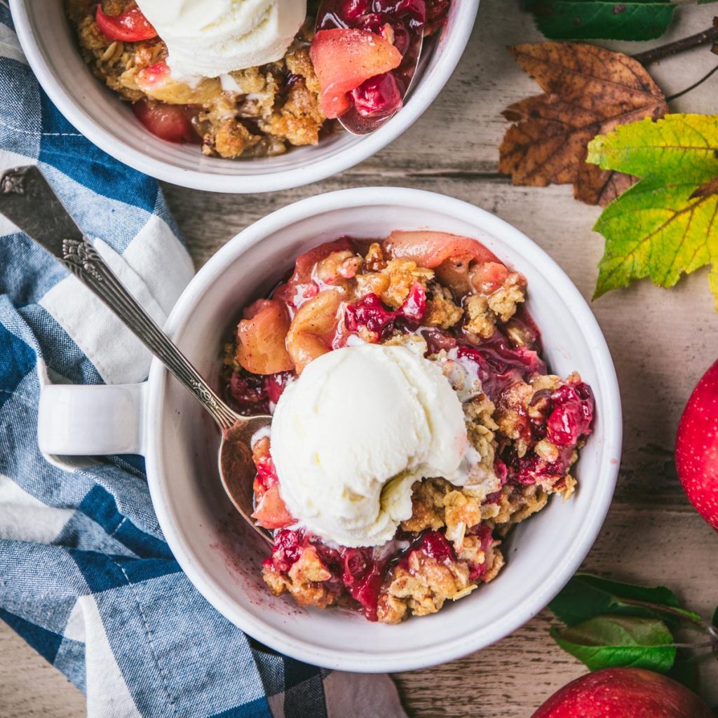 Square overhead shot of cranberry apple crisp in a white bowl with ice cream