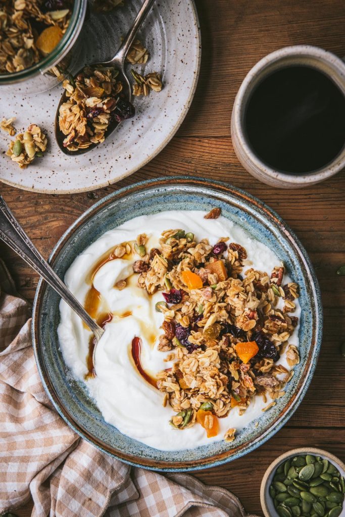 Overhead shot of a bowl of homemade granola with yogurt and maple syrup on a breakfast table