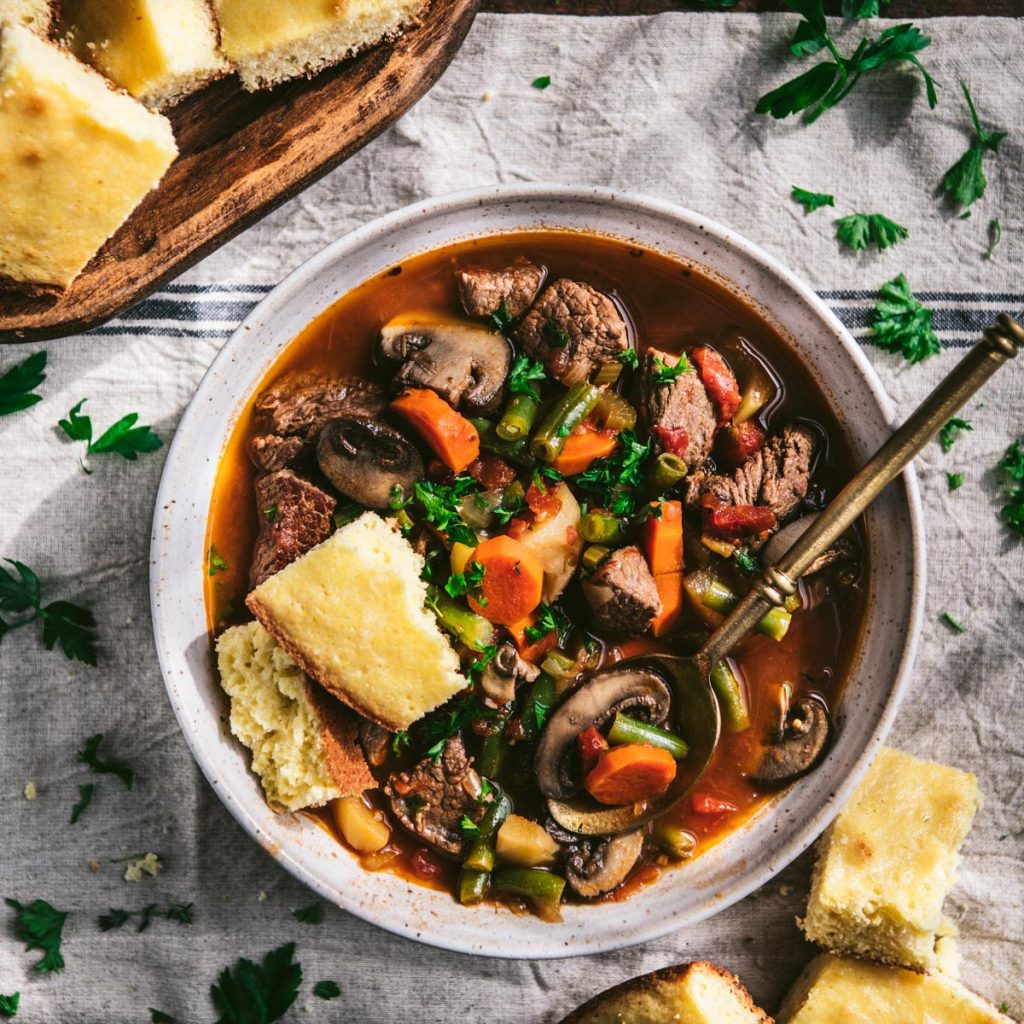Square overhead shot of a bowl of vegetable and beef soup with cornbread on the side