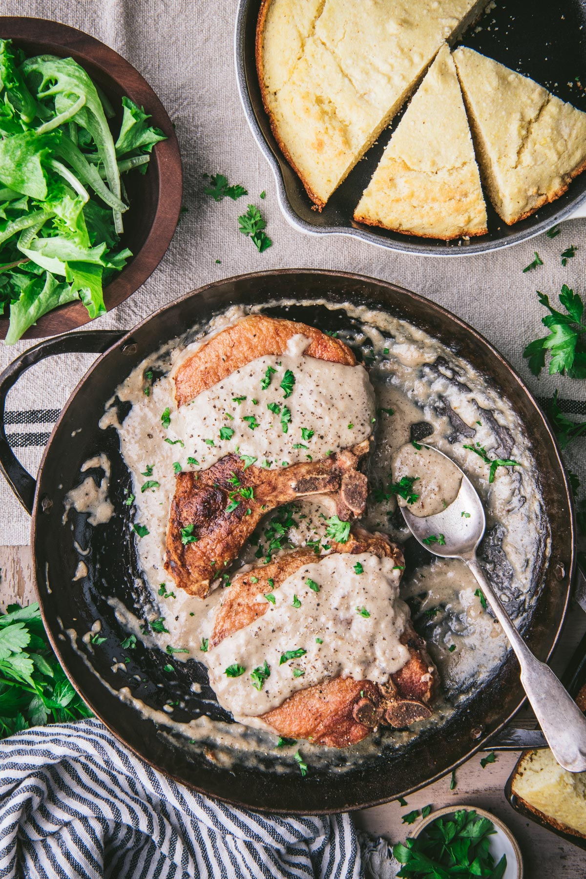 Pork chops and gravy in a cast iron skillet on a dinner table with a side of cornbread and salad