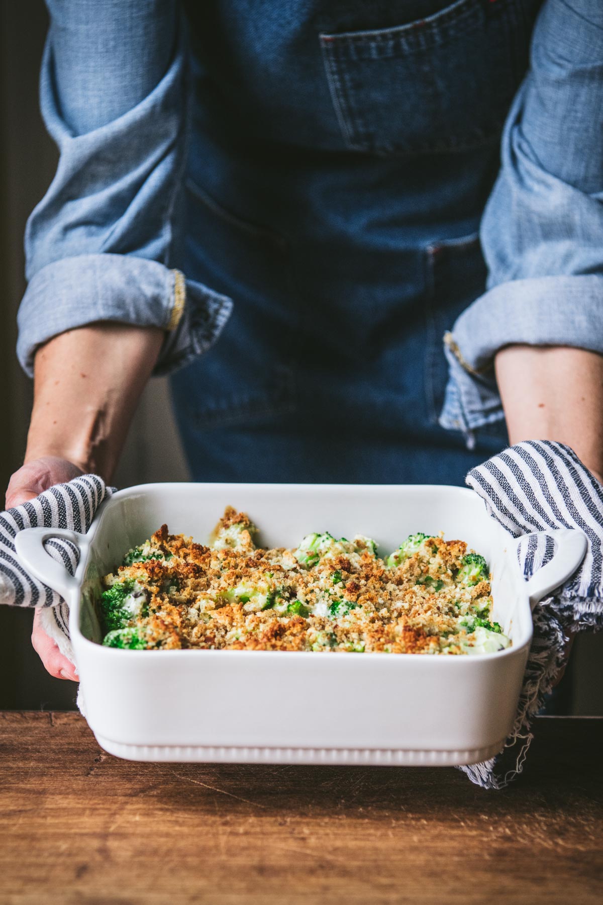 Hands placing a broccoli casserole in a white dish on a wooden table