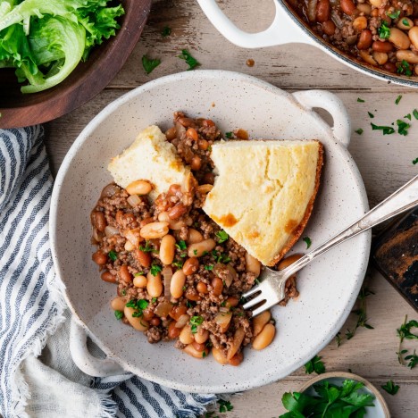 Square overhead shot of a bowl of ground beef and baked bean casserole with cornbread on a dinner table