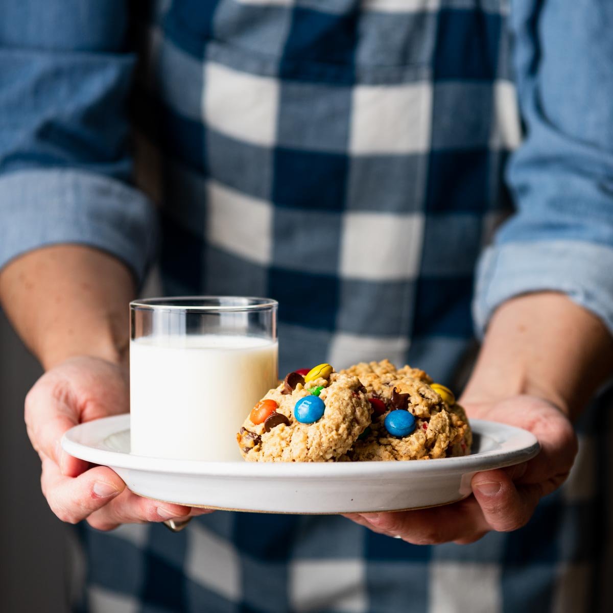 Square front shot of hands serving a white plate of monster cookies and milk