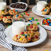 Square image of two white plates full of monster cookies on a table with milk, M&Ms, and chocolate chips
