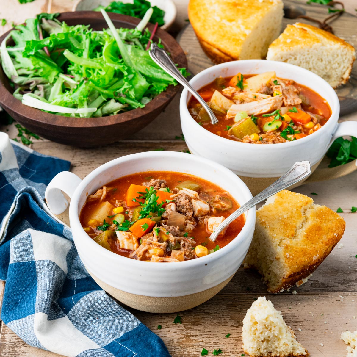 Square side shot of two bowls of burgoo on a table with salad and cornbread