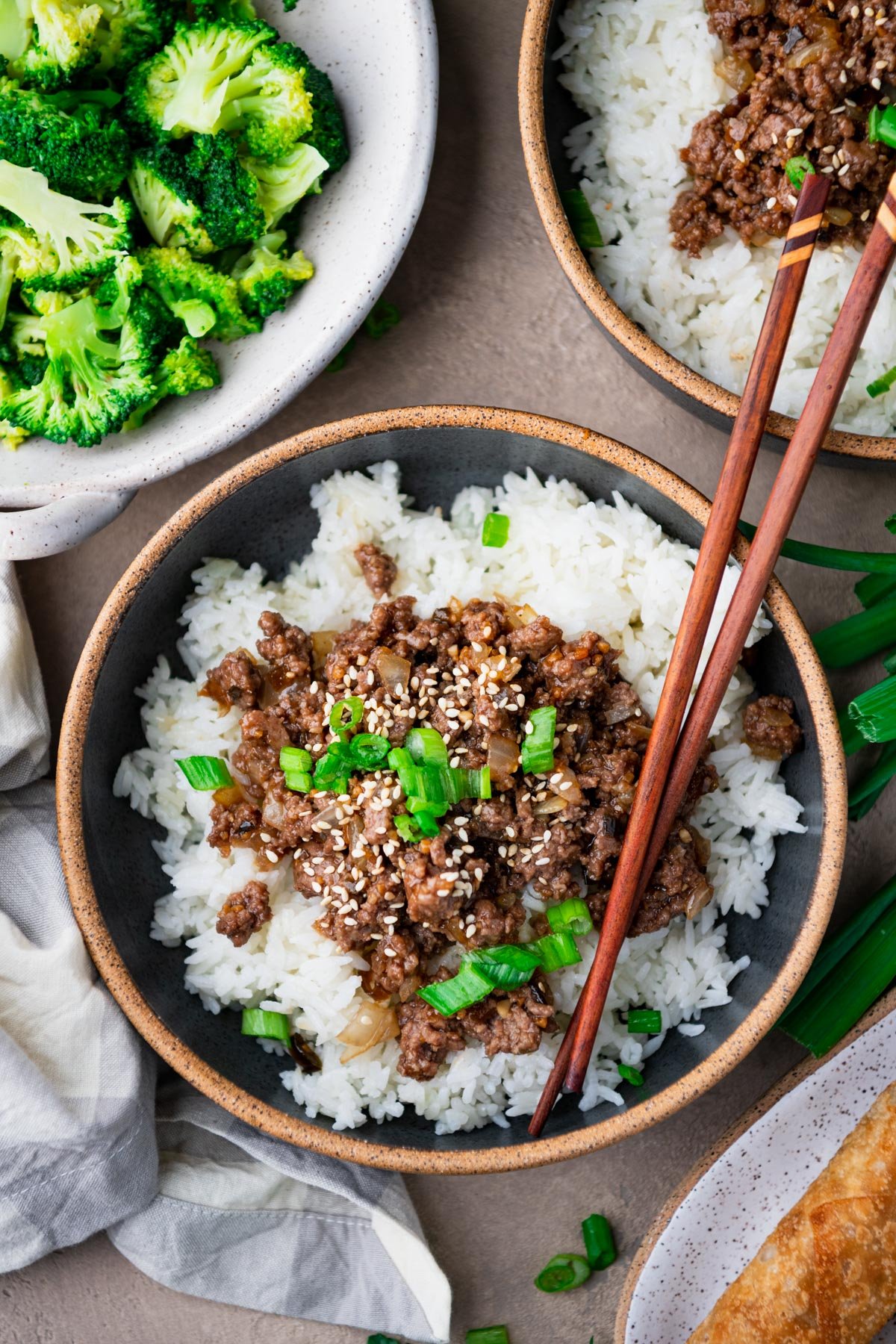 Close overhead image of a bowl of ground beef teriyaki over rice on a table with broccoli and egg rolls.