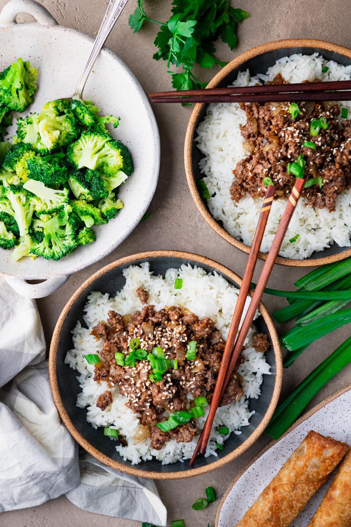Overhead image of two bowls of ground beef teriyaki recipe on a table with a side of broccoli and egg rolls.
