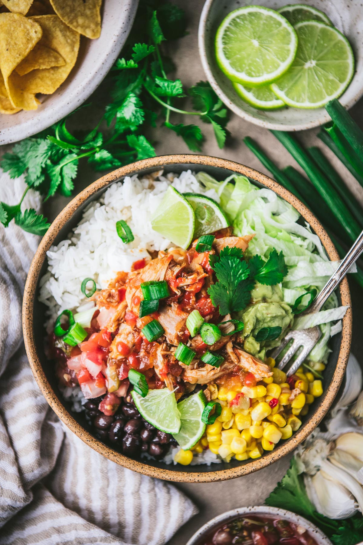 Overhead shot of a fork in a bowl of crock pot chipotle chicken with limes, corn, beans, guacamole, and pico de gallo.
