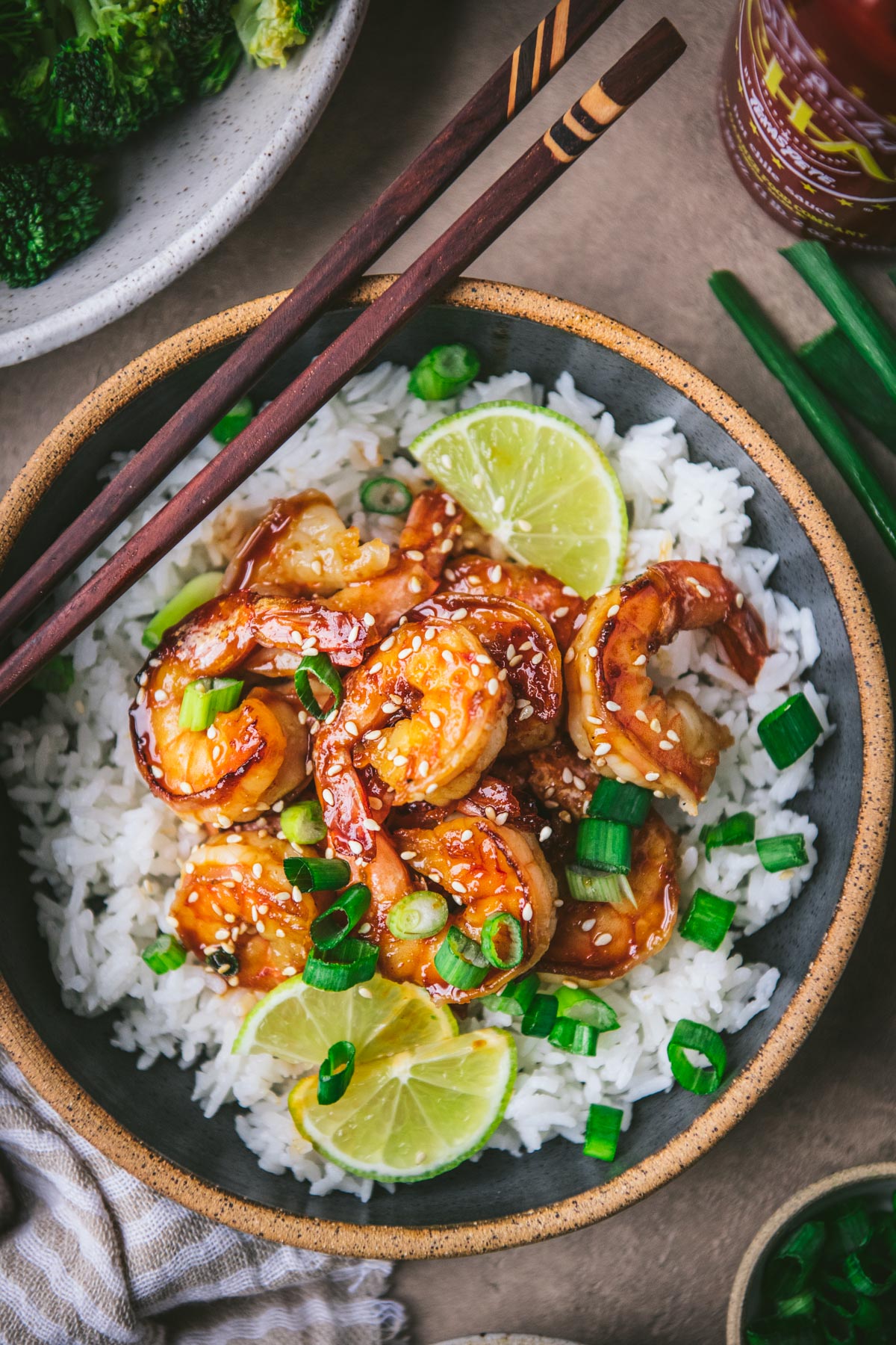 Close overhead image of a bowl of honey sriracha shrimp garnished with sesame seeds, limes, and green onions.