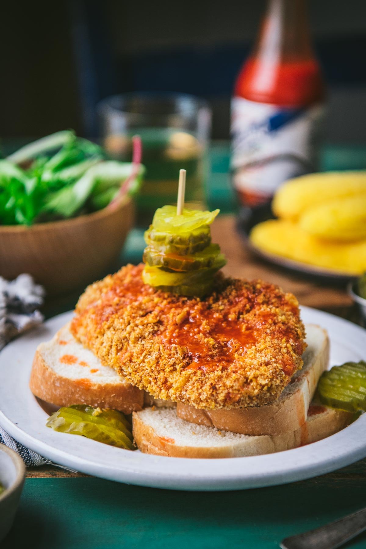 Side shot of a baked nashville hot chicken recipe served on a white plate with white bread and sliced pickles.