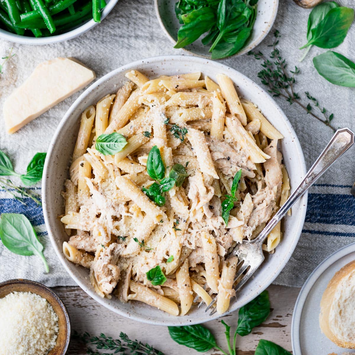 Square overhead shot of a bowl of crock pot garlic parmesan chicken pasta on a table with fresh basil on top