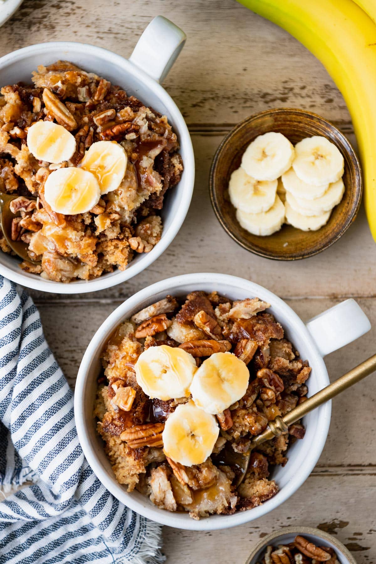 Two bowls of dump cake with bananas on a table with extra sliced bananas.