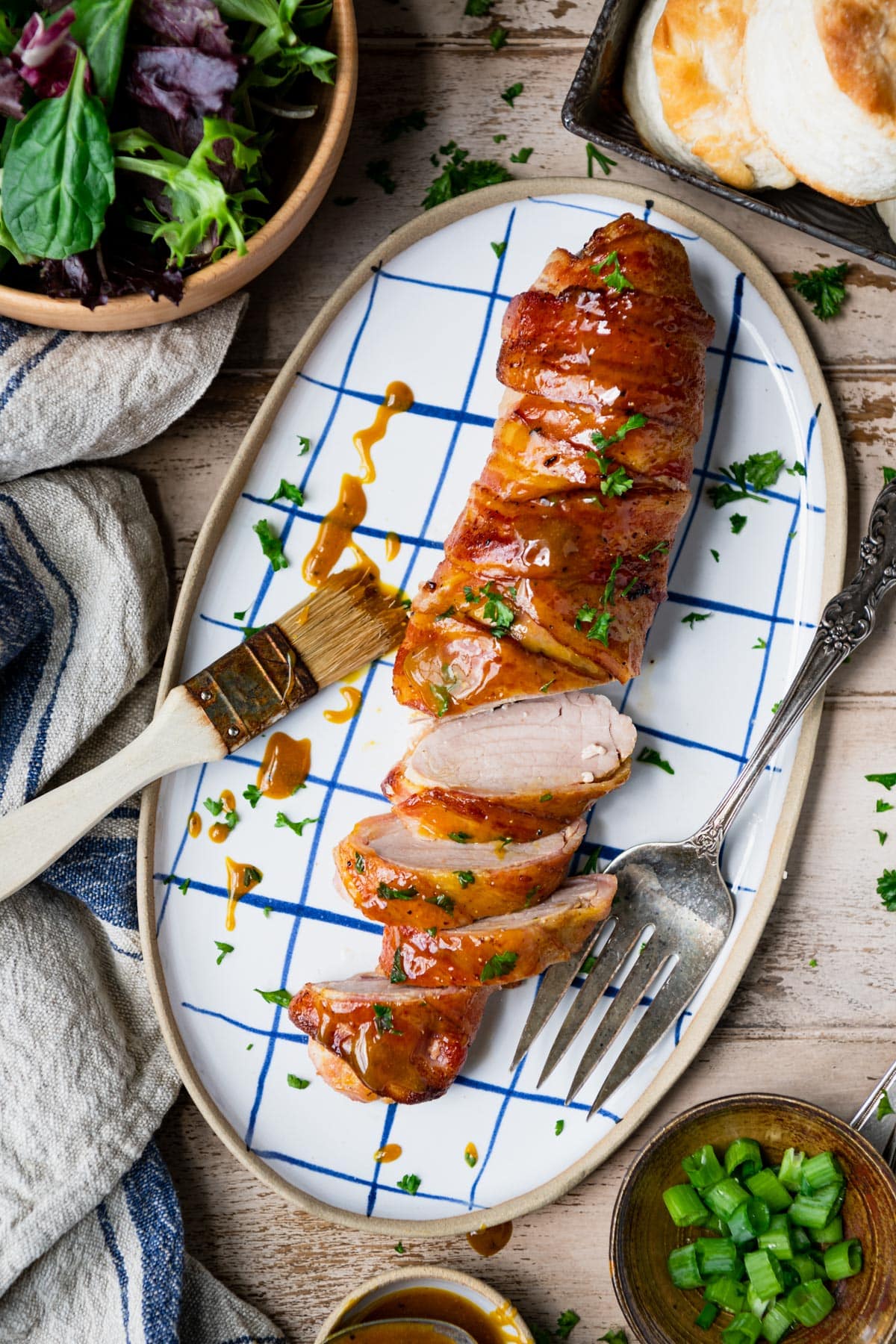 Bacon wrapped pork tenderloin with brown sugar and mustard served on a blue and white tray with fresh parsley.