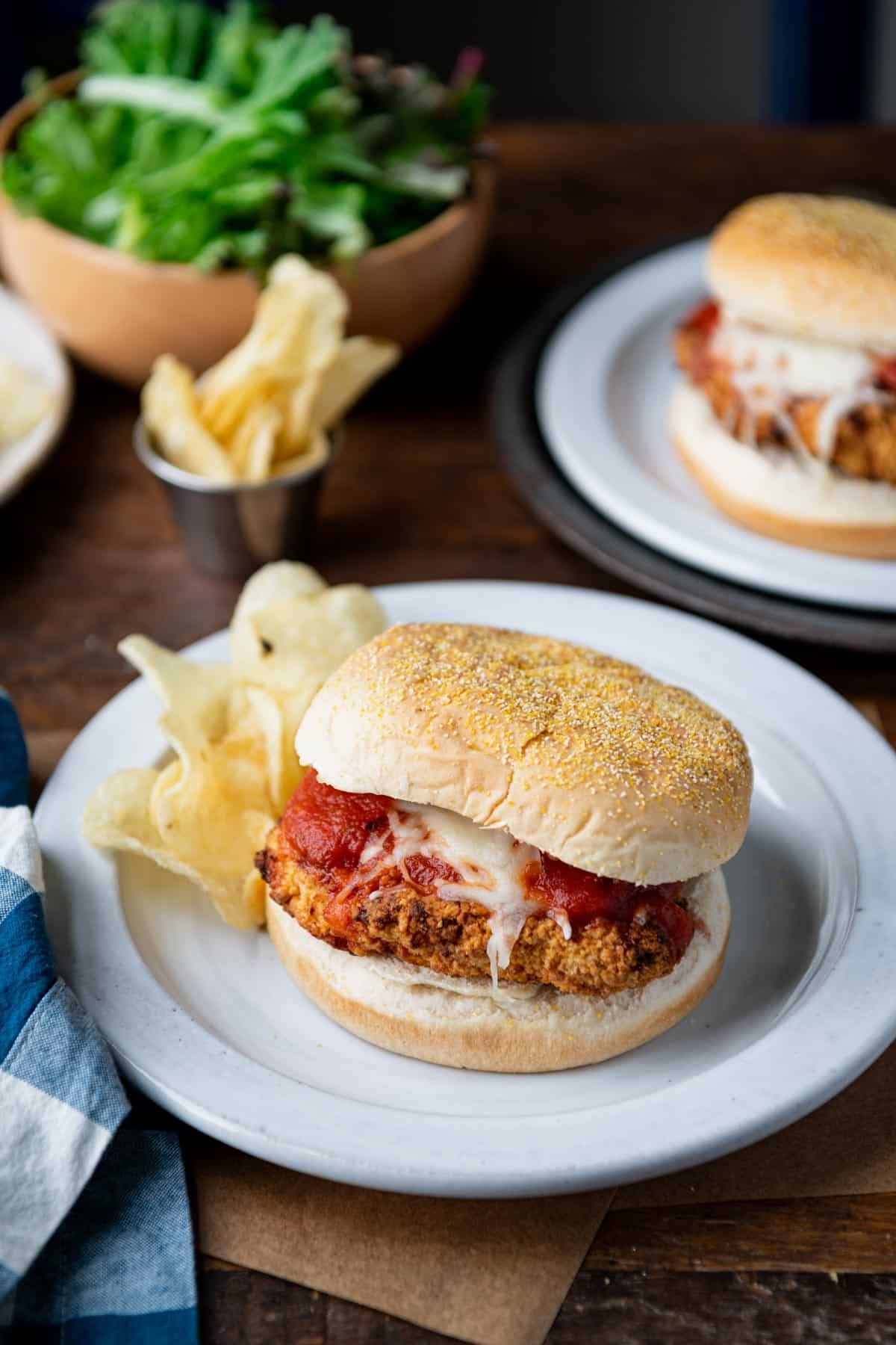 Two chicken parm sandwiches on white plates on a wooden table.