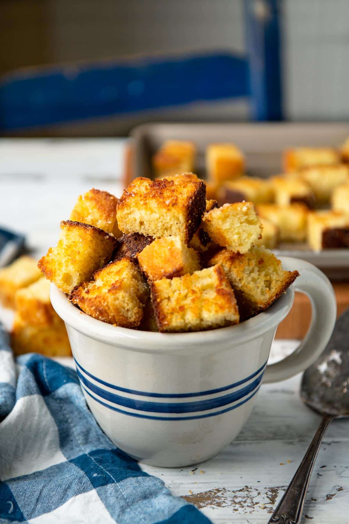 Homemade cornbread croutons stacked in a blue and white mug on a table.