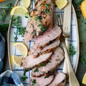 Overhead shot of marinated grilled pork tenderloin on a blue and white check platter with fresh herbs and lemon slices.