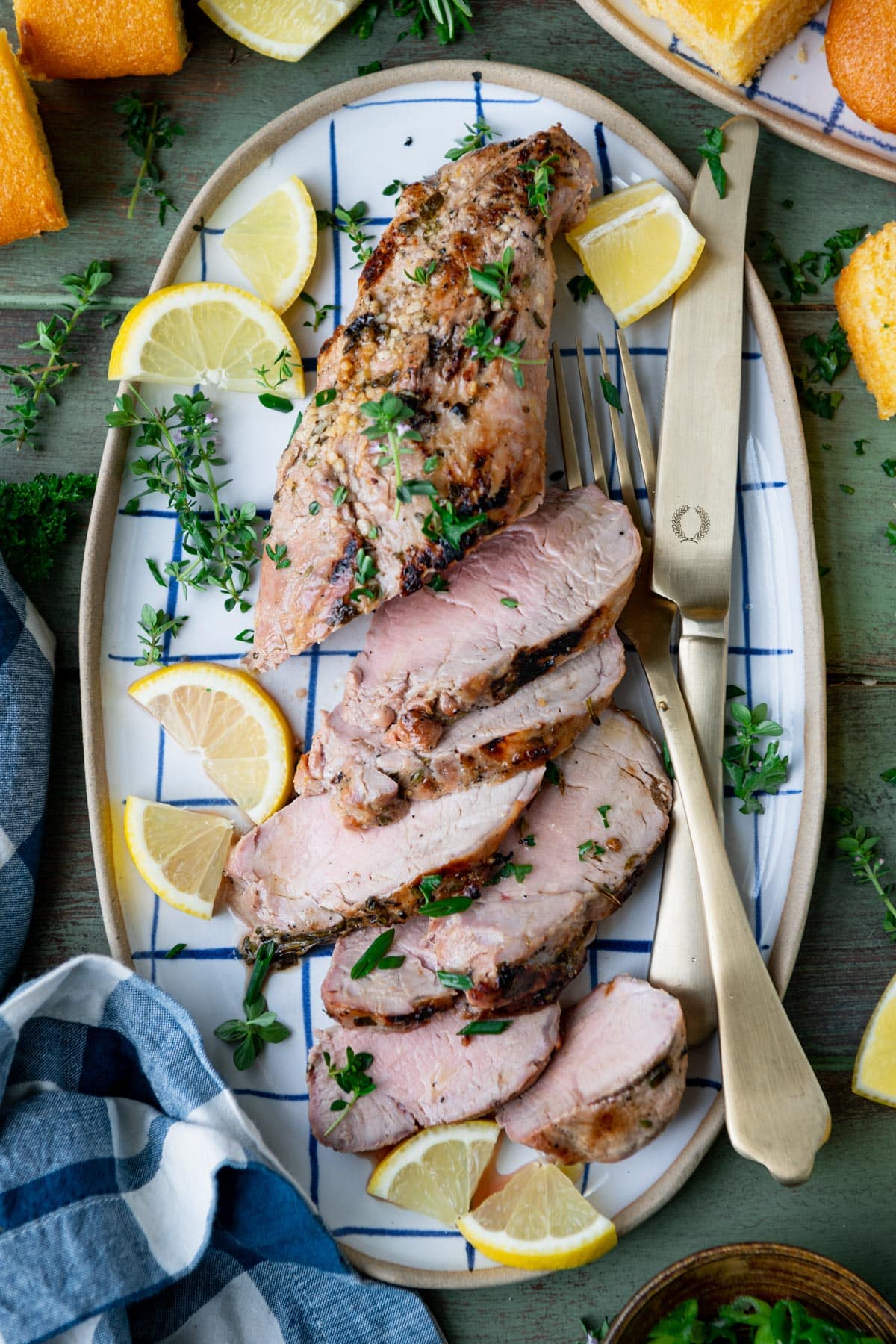 Overhead shot of marinated grilled pork tenderloin on a blue and white check platter with fresh herbs and lemon slices.